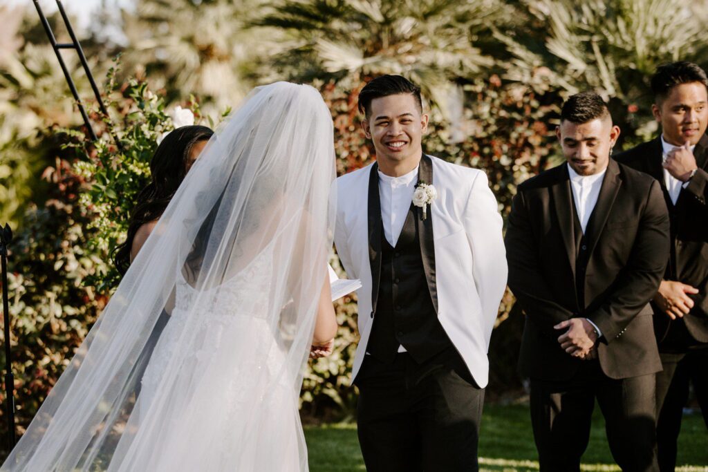 Groom in white jacket laughing during wedding ceremony. 