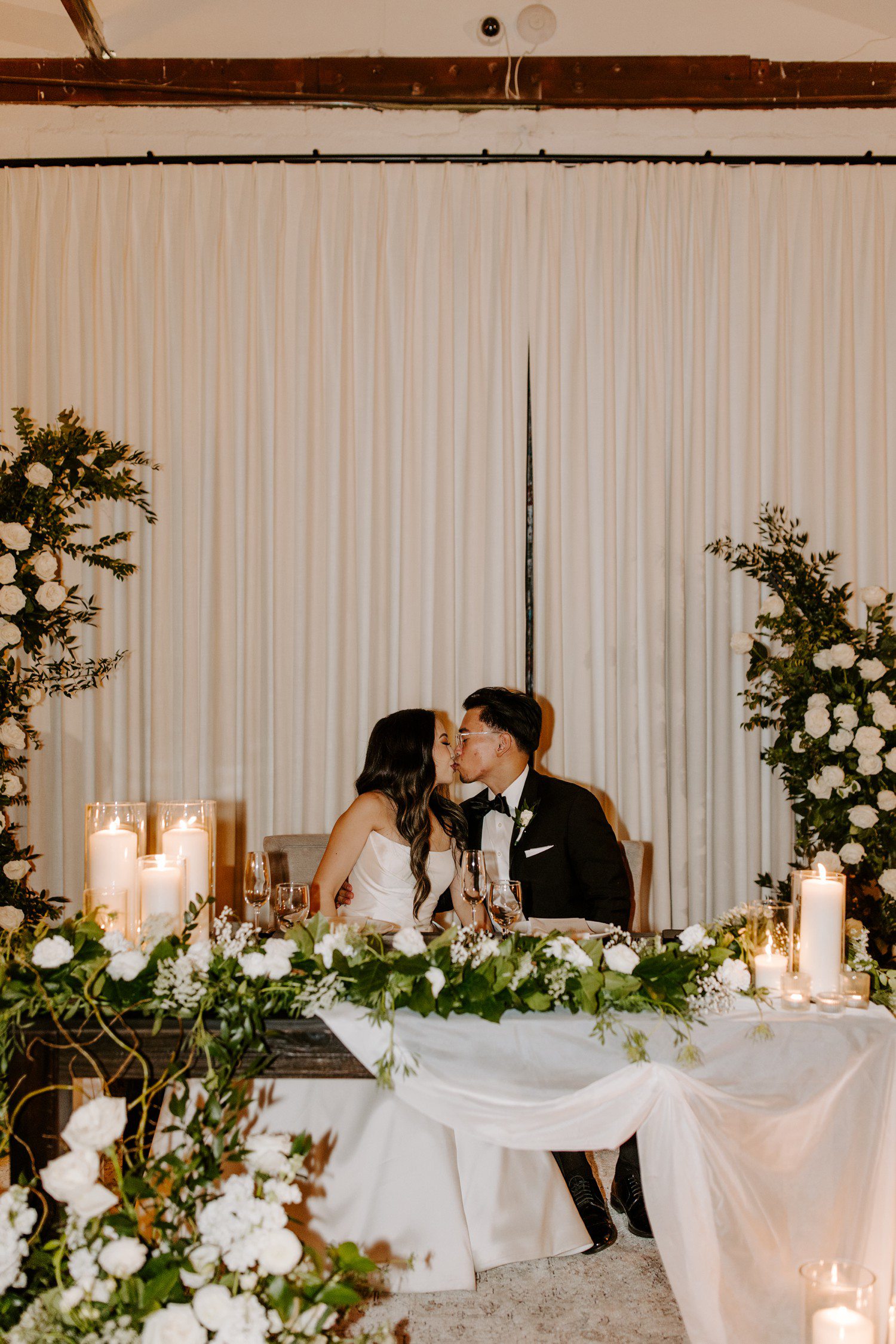 Bride and groom kiss at reception table.