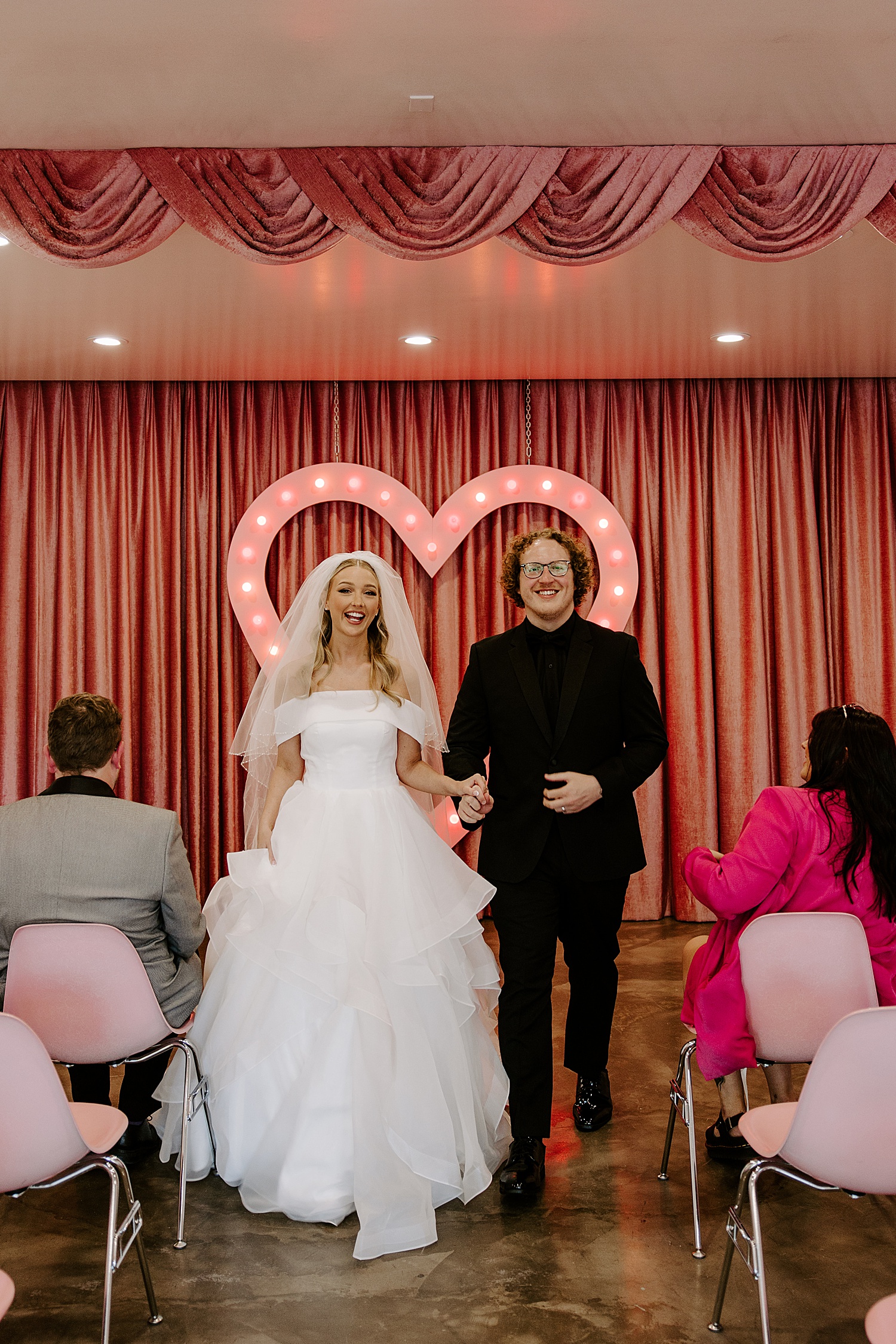 newlyweds exit ceremony in front of pink light heart by Katelyn Faye Photography