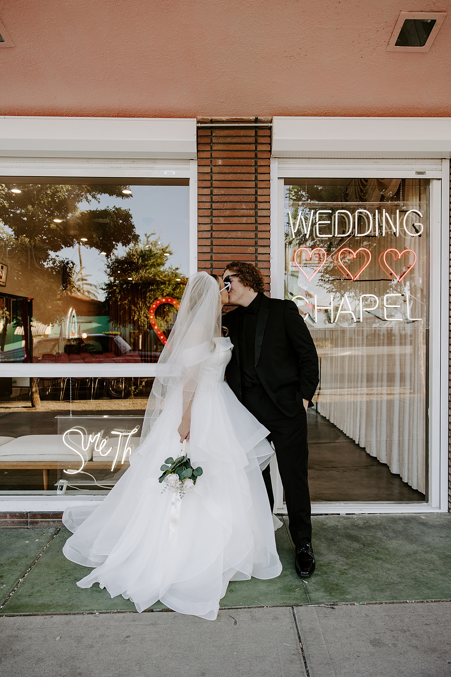 bride in white sunglasses kissing in front of glass windows by Katelyn Faye Photography