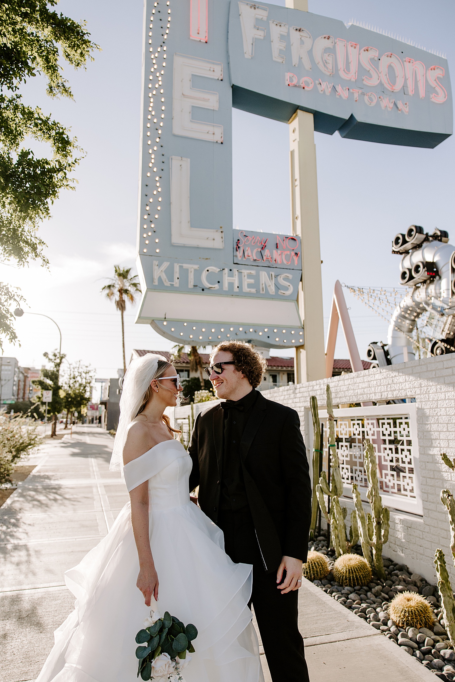 man in black suit smiles at woman in white gown under big blue sign by Las Vegas elopement photographer