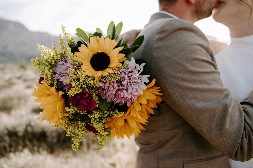 woman hangs hand holding wedding florals over man's should while they kiss by Katelyn Faye Photography