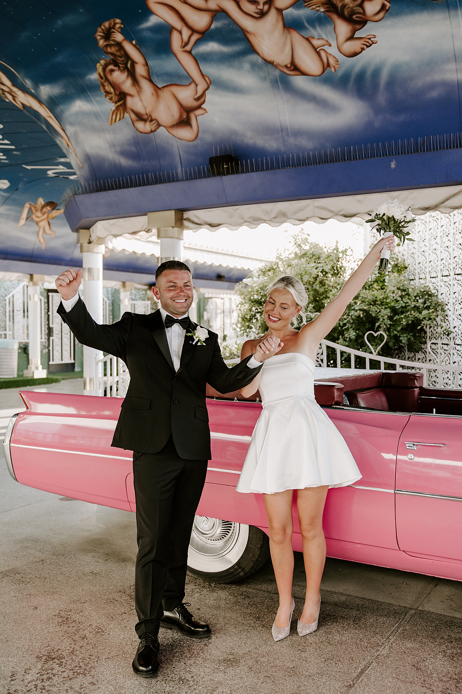 newlyweds throw hands in the air next to pink corvette in the Tunnel of Love at the LWC