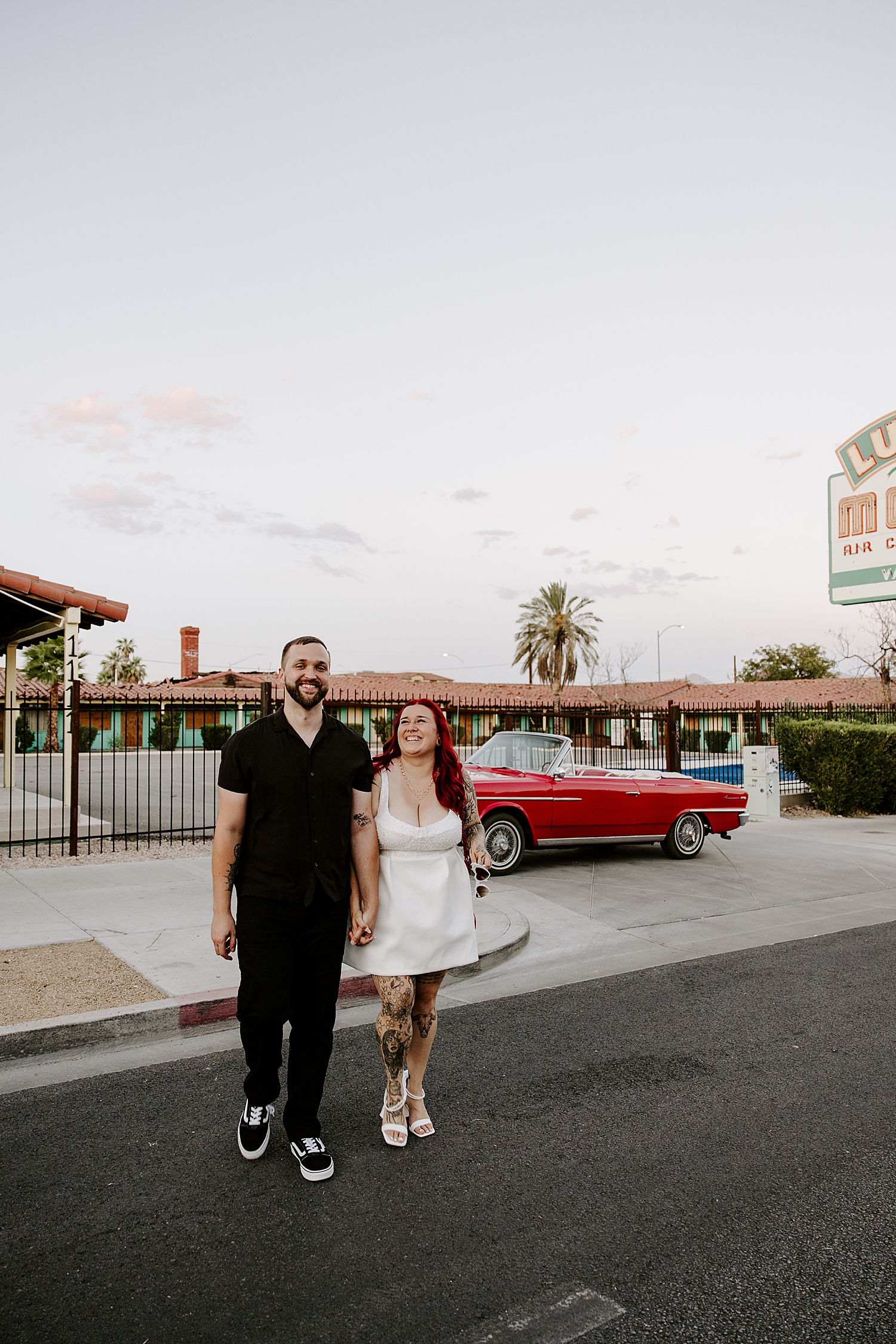 man in all black, and woman in white walk across the street from vintage car