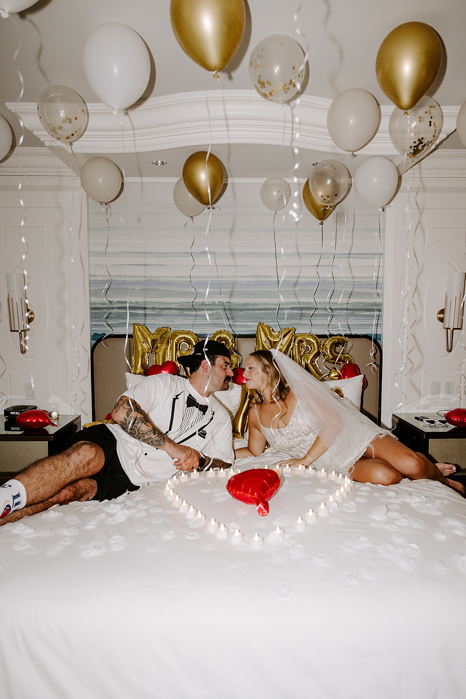 bride and groom lie across bed under gold balloons by Katelyn Faye Photography